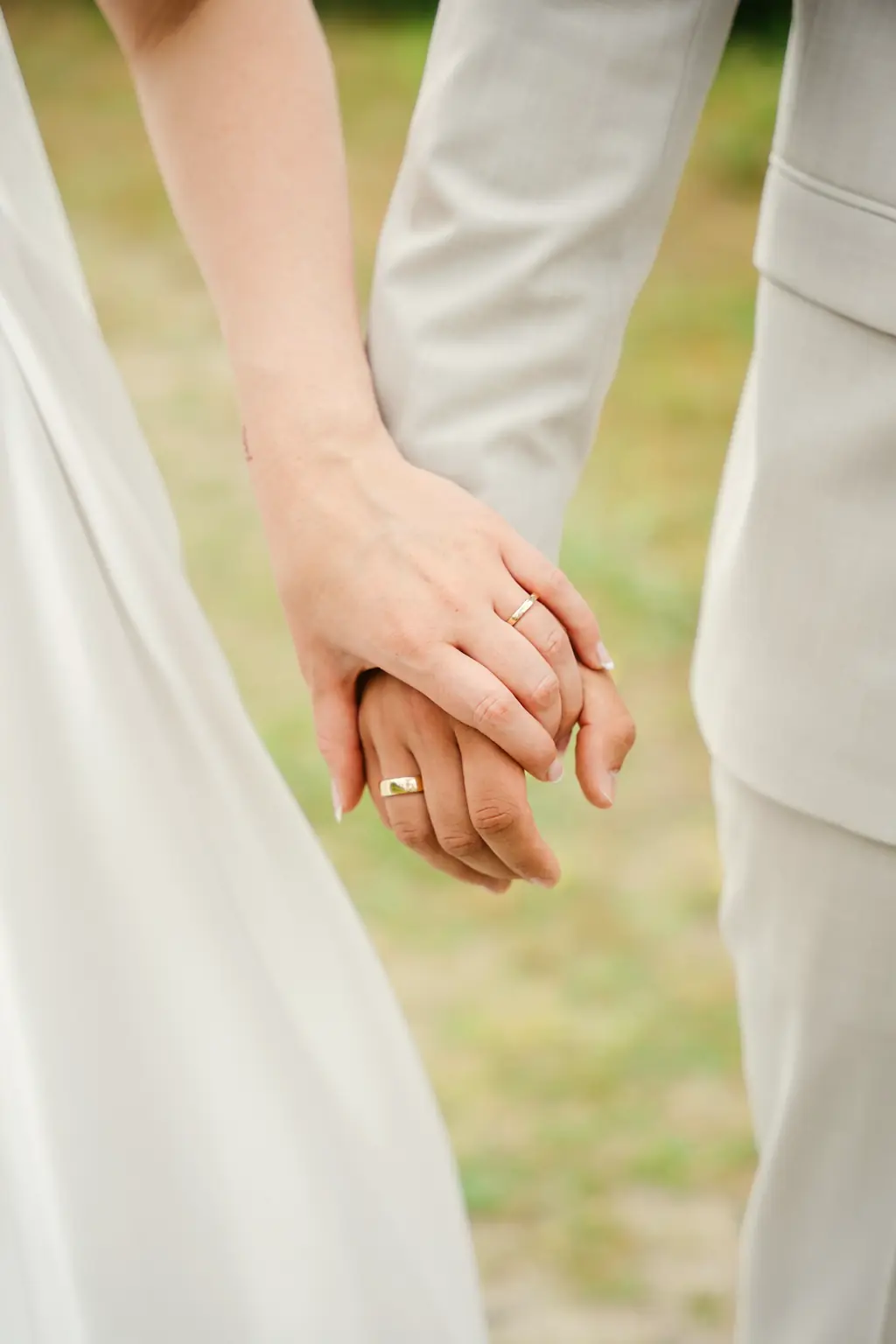 The couple's hands, fingers laced, both rings showing against a white dress and a pale suit.