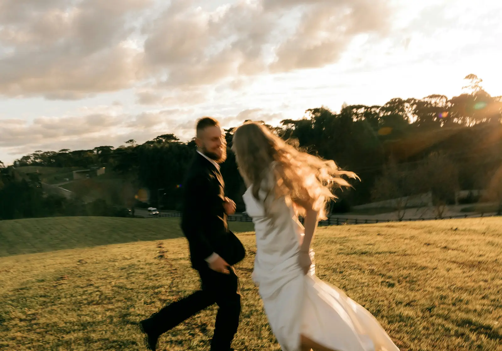 A bride and groom running hand in hand across a hillside at golden hour, her hair and dress flying.