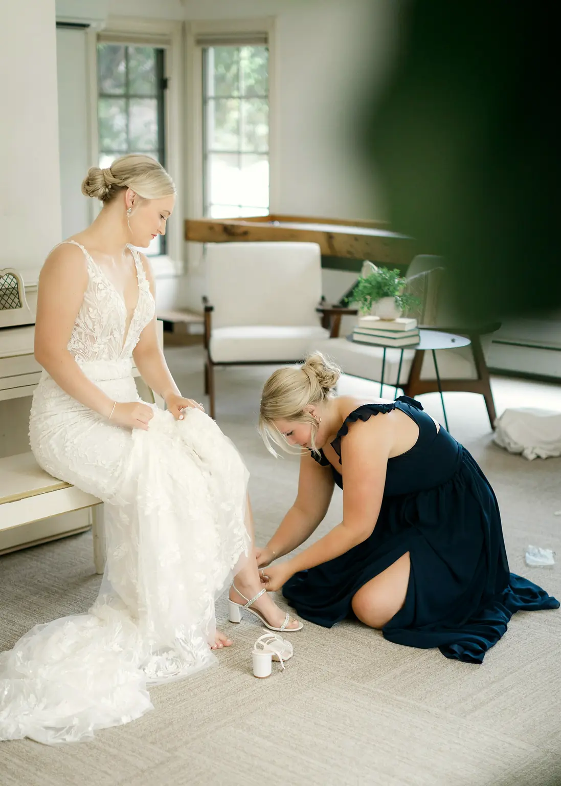 A bridesmaid kneeling to buckle the bride's shoe in a bright sitting room.