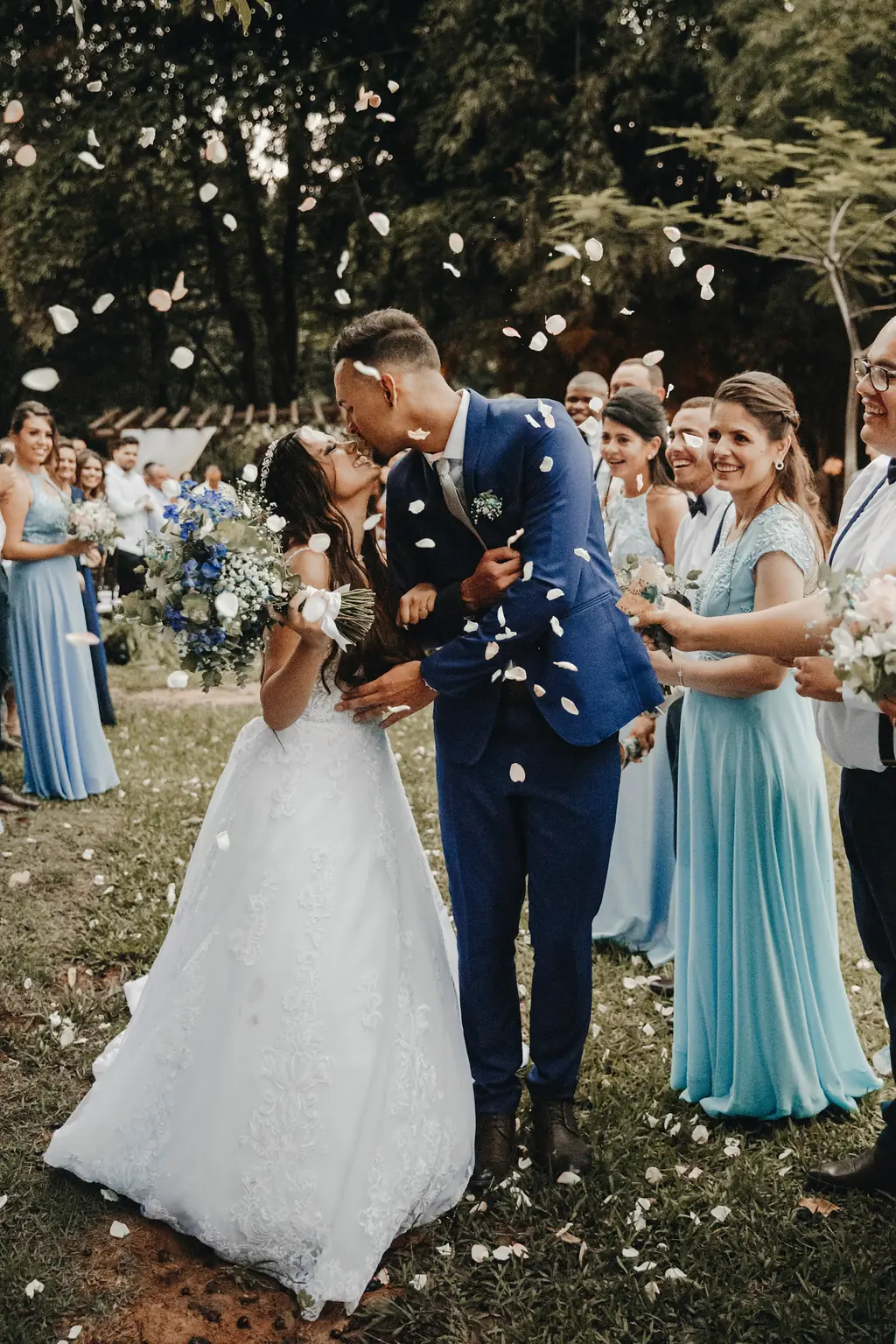 The couple kissing in a shower of white petals, bridesmaids in blue laughing around them.