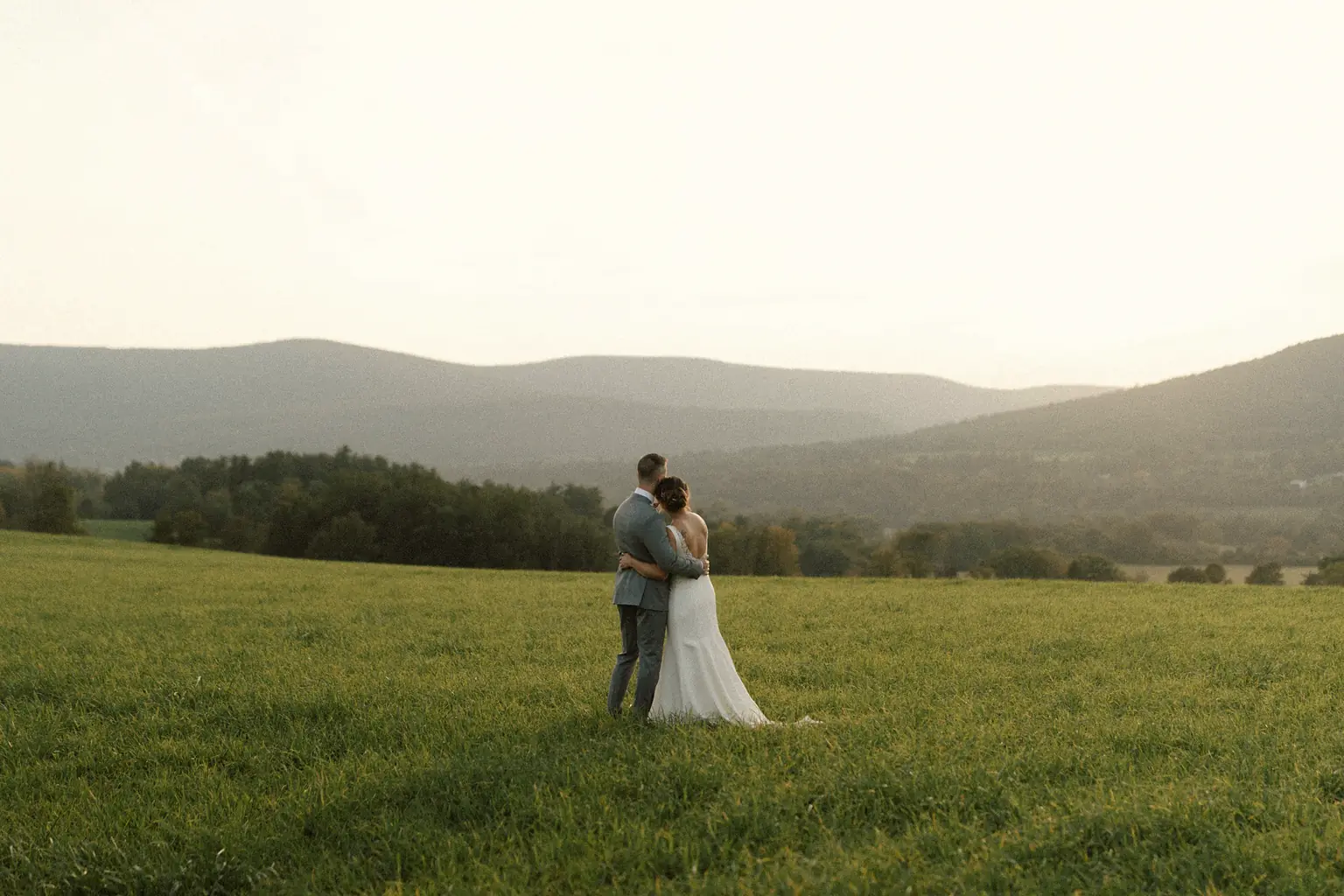 A couple holding each other in a wide green field, hills fading into haze behind them.