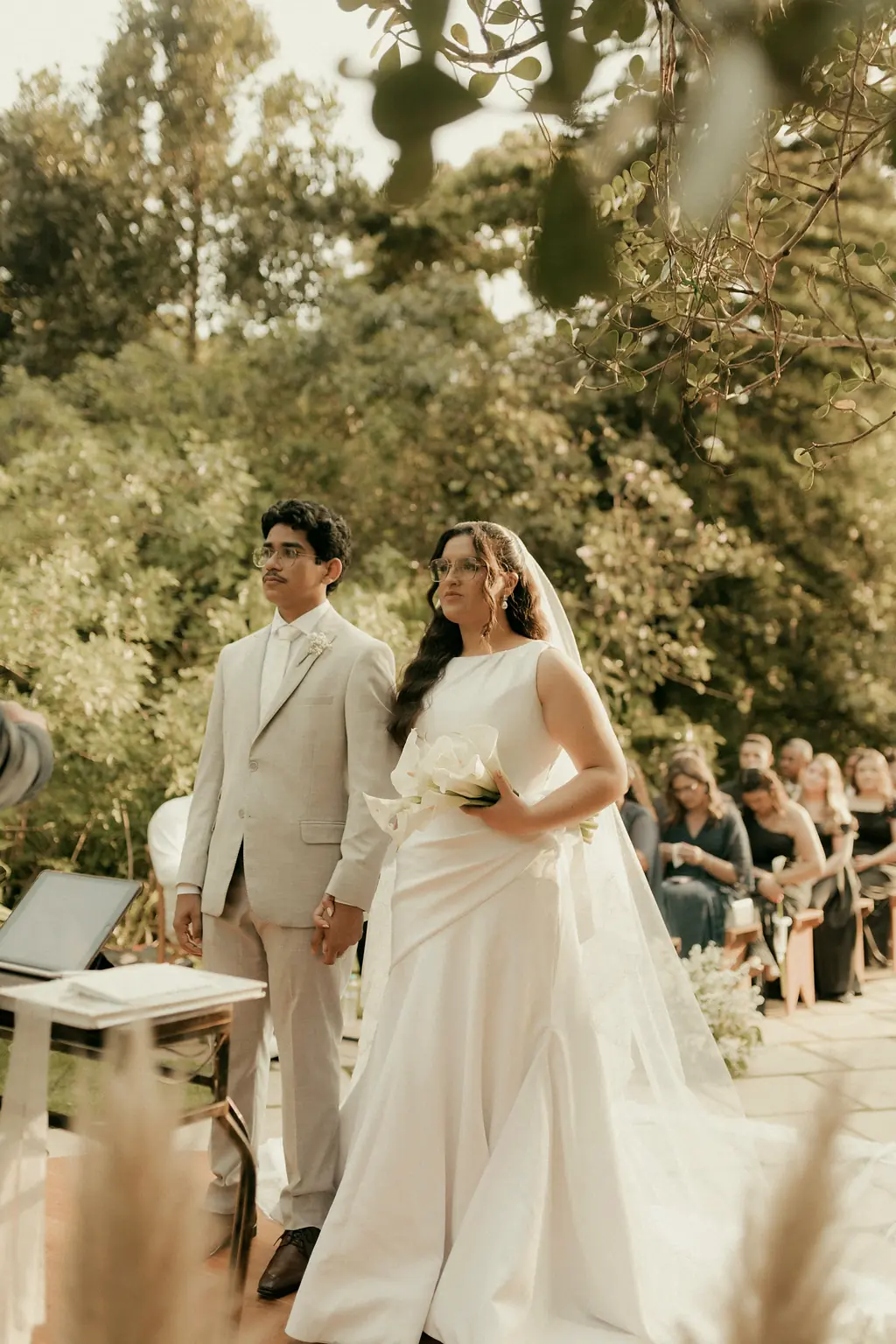 A bride and groom standing at a garden ceremony among tall trees, guests seated behind them.