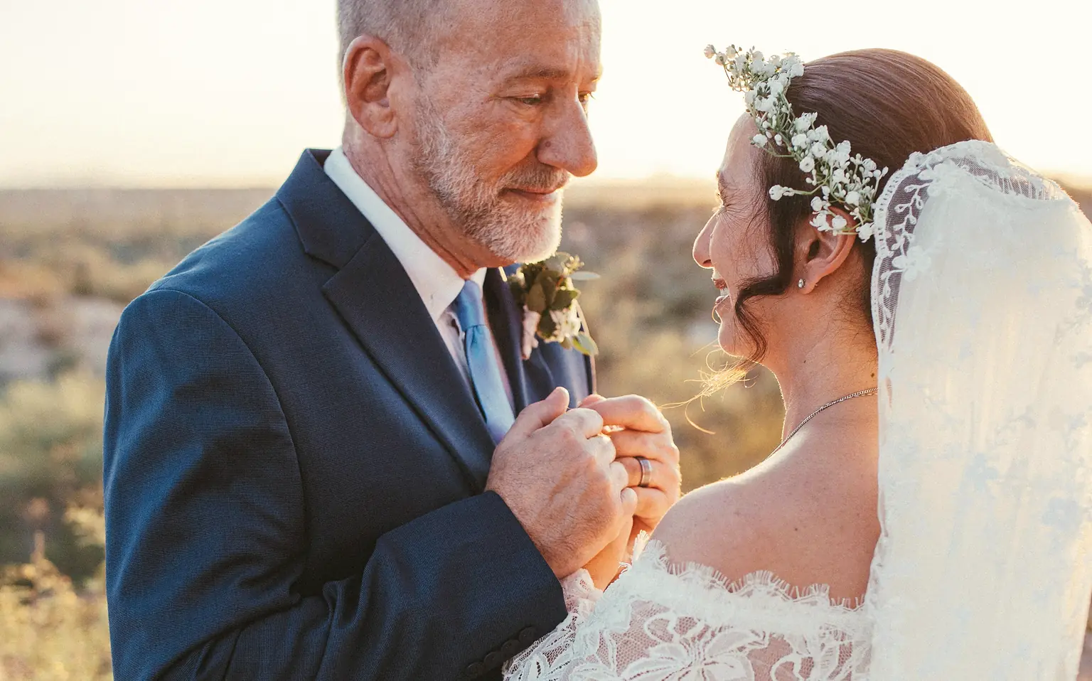 An older groom holding the bride's hands, both smiling, in warm evening light.