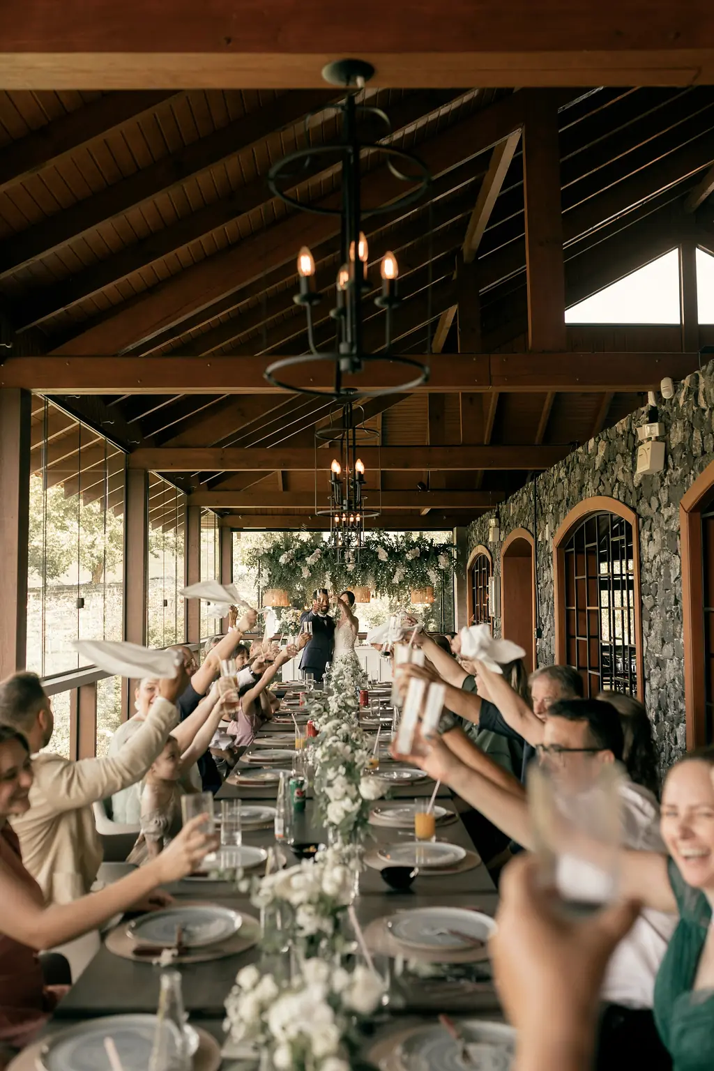 Guests along a long table under a timber roof, glasses raised in a toast.