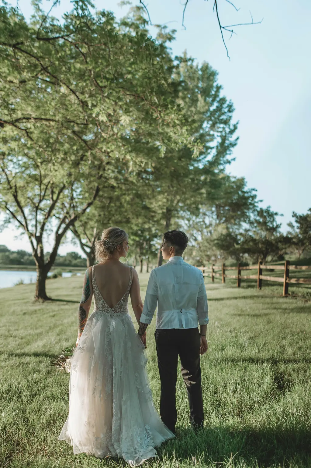 The couple walking hand in hand along a row of trees by a lake, seen from behind.